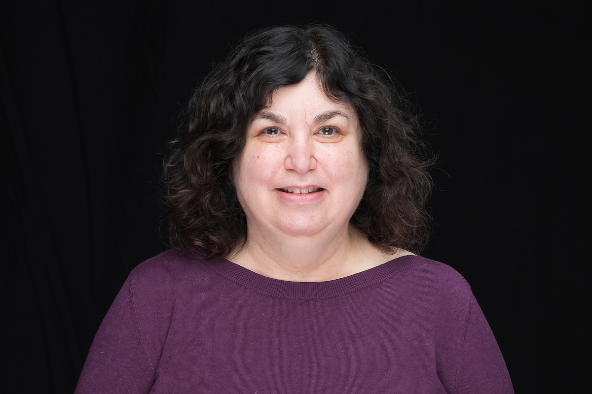 Roni Reiter-Palmon stands in front of a black background. She has dark hair and is wearing a purple shirt. 