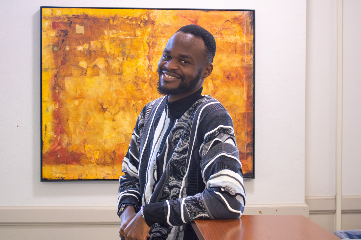 A man wearing a patterned sweater leans on a desk and smiles warmly. Behind him is an abstract orange and yellow painting on a white wall.
