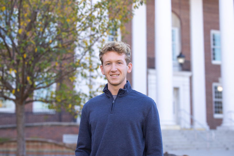 Simon smiles in front of a building with tall pillars and autumn leaves. He is wearing a navy sweater.